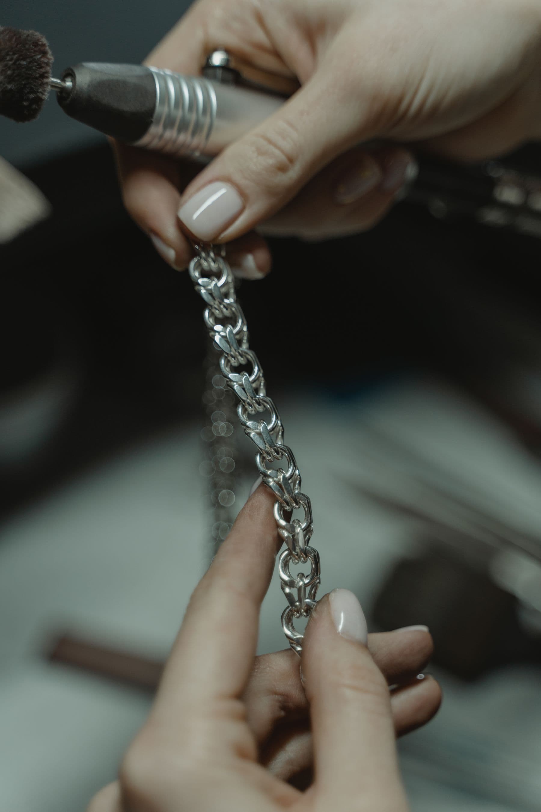 A silver chain held up to a rotary tool at a jeweler's bench.