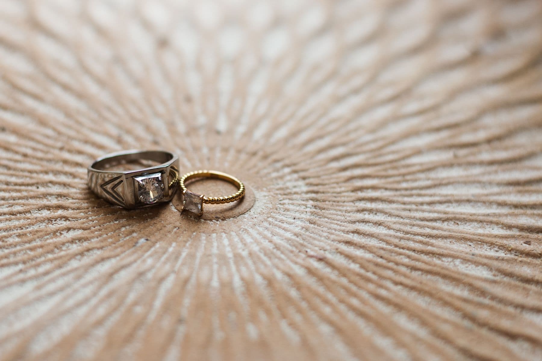 A silver ring with a square stone beside a thin gold ring on carved wood.